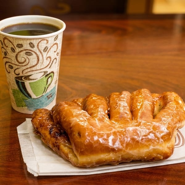 A glazed bear claw pastry on a white napkin next to a cup of coffee on a wooden table.