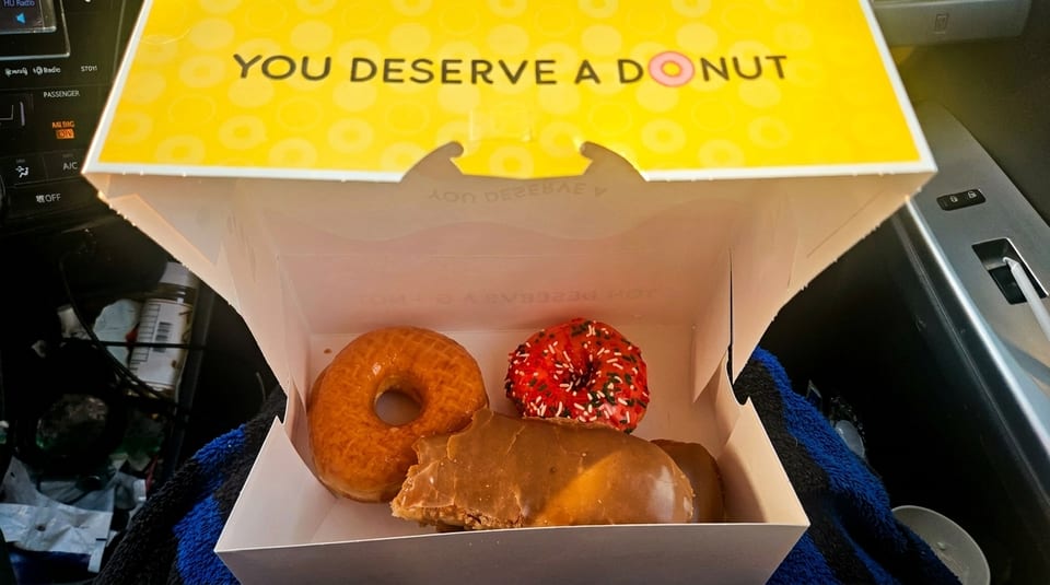 An open donut box with "YOU DESERVE A DONUT" on the lid, containing three assorted donuts.