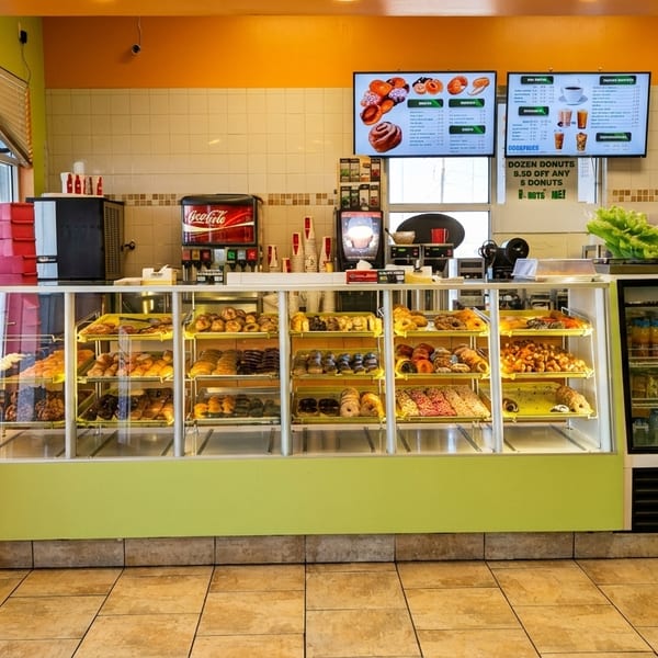 A brightly lit donut shop interior with a long display case filled with various pastries and drinks.
