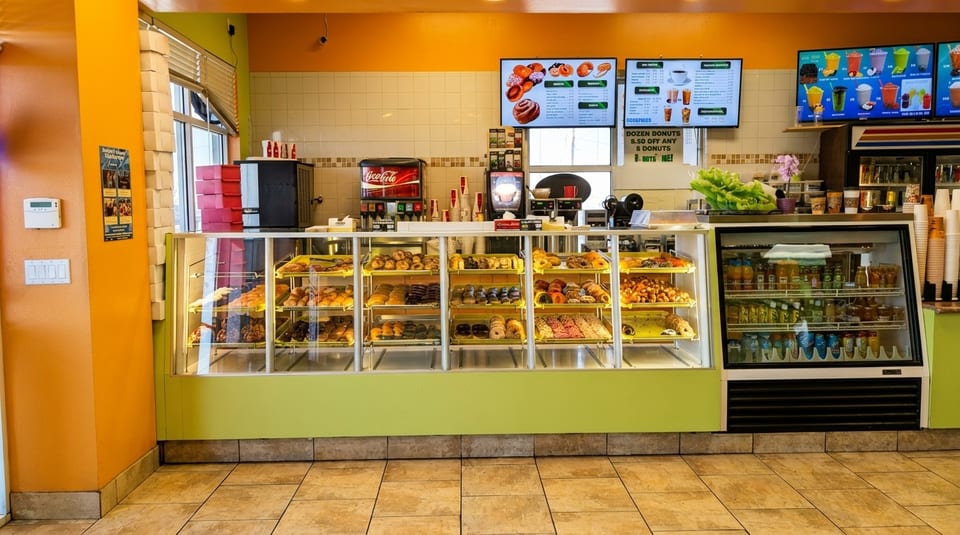 A brightly lit donut shop interior with a long display case filled with various pastries and drinks.