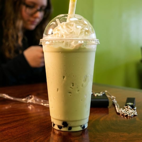 Green boba tea with whipped cream and a straw on a wooden table, with a person in the background.
