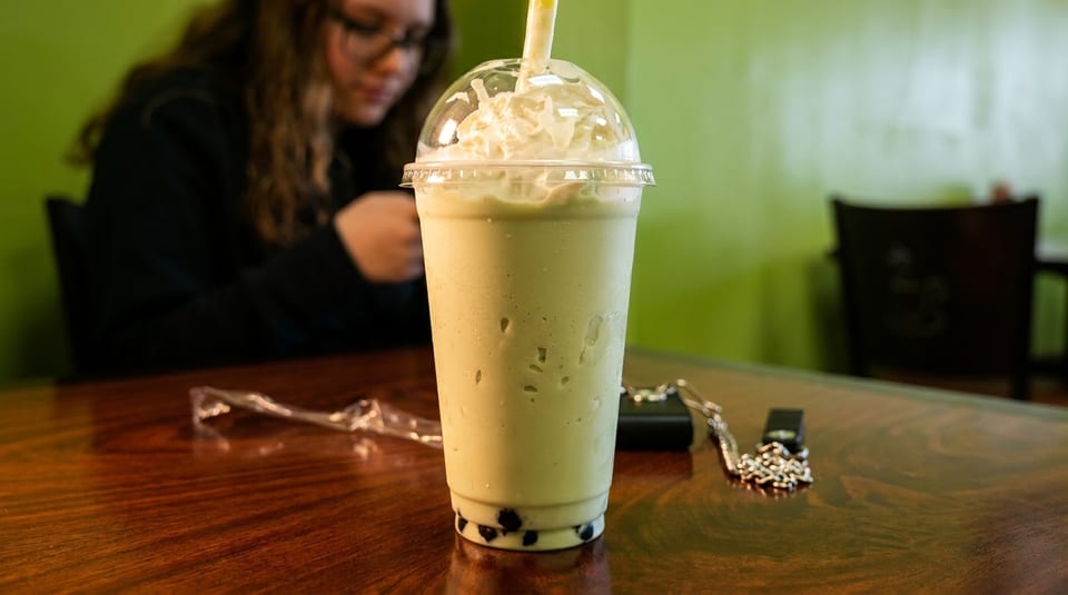 Green boba tea with whipped cream and a straw on a wooden table, with a person in the background.